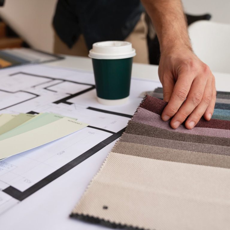 Beispielbild zur Planung Innenausbau Farbmuster und Pläne auf einem Tisch, Hand hält einen Streifen, daneben ein Kaffeebecher.