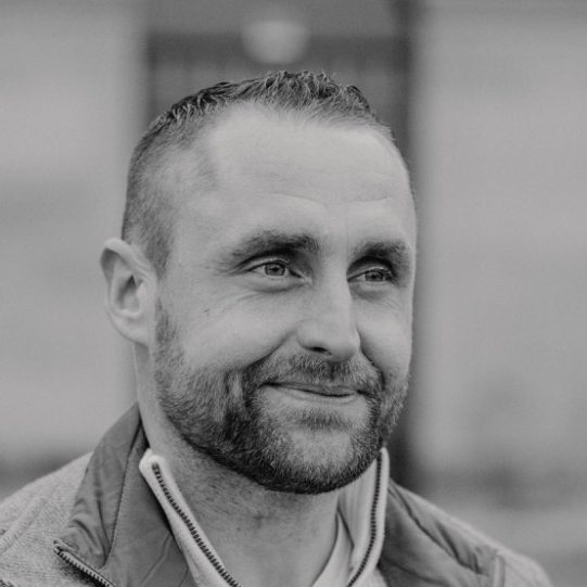 Brian Krüger mit einem Lächeln, trägt eine Jacke, unscharfer Hintergrund.