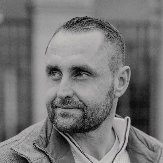 Brian Krüger - Inhaber Porträt von Brian Krüger mit Kurzhaarschnitt und Bart, lächelnd in Schwarz-Weiß.