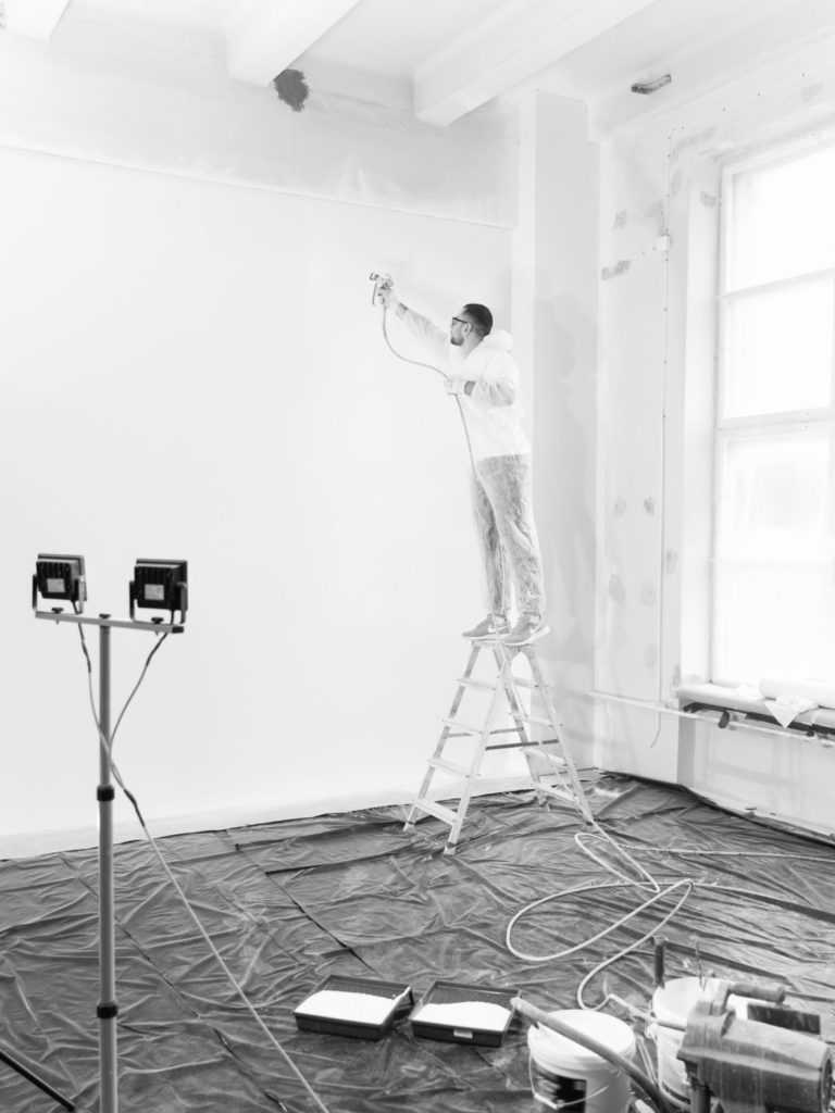 Maler streicht eine Wand Mann auf Leiter streicht weiße Wand in leeren Raum mit Arbeitsmaterialien am Boden.