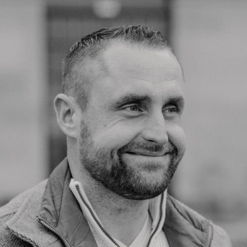 Malermeister Brian Krüger Lächelnder Brian Krüger, hellem Oberteil und grauer Jacke, in unscharfer Umgebung.