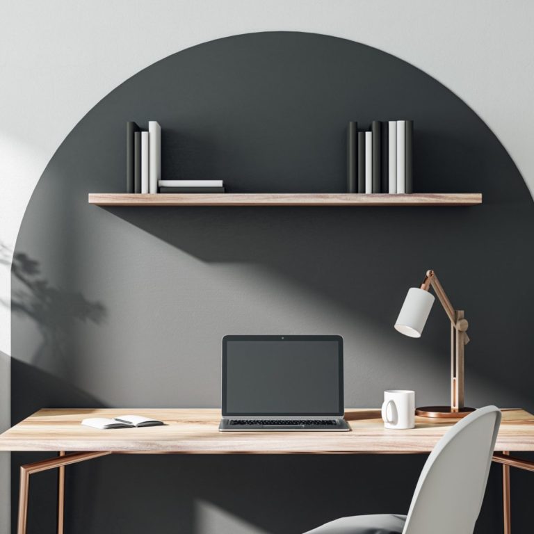 Ein moderner Schreibtisch mit Laptop, Schreibtischlampe und Wandregal über einem grauen Hintergrund.