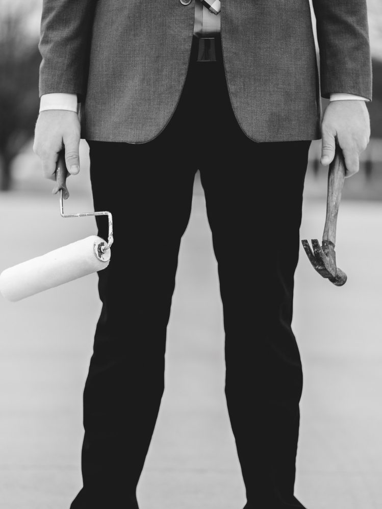 Mann in Anzug mit Farbrolle und Hammer in den Händen, steht auf einem Gehweg.