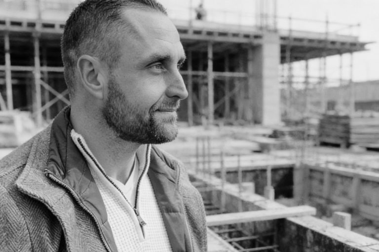 Brian Krüger auf der Baustelle Brian Krüger auf einer Baustelle, lächelnd, mit einem Baugerüst im Hintergrund.